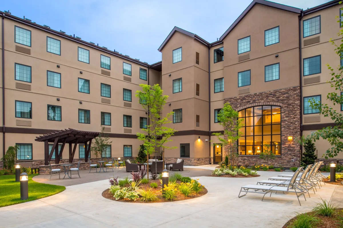 This image displays the modern facade of Staybridge Hillsboro North, set against a landscaped garden with a seating area under a pergola and lounge chairs. The scene is tranquil and inviting, with sunset lighting enhancing the atmosphere. The setup suggests a place where families can gather, relax, or enjoy the outdoors together.