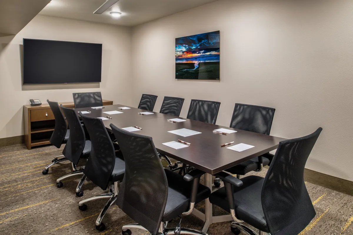 This image features a spacious interior conference room setup with a large table, ergonomic chairs, a mounted television, and tasteful landscape artwork. The environment is modern and professional, yet comfortable, echoing an atmosphere that supports both productivity and relaxation.