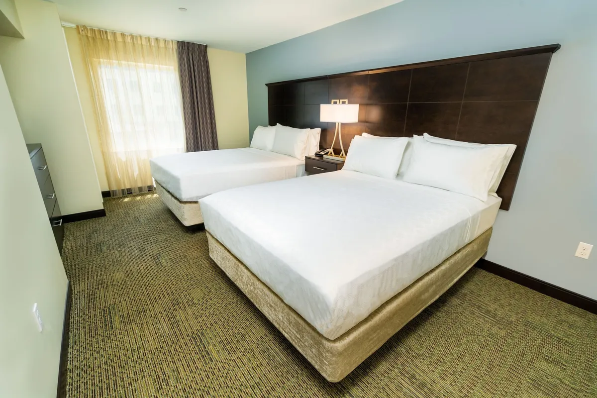 This image features two inviting double beds with crisp linens, a nightstand with a warm lamp, and a large window filling the room with natural light. The setting is calm, restful, and designed for comfort, perfectly reflecting the suite’s atmosphere.