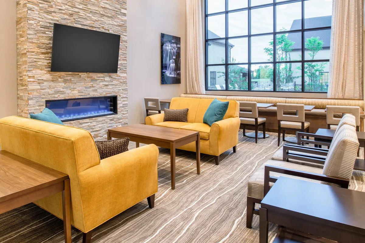 A cozy interior living area with inviting yellow sofas, large windows allowing natural light, a decorative stone wall, a fireplace, and artwork. The layout connects seamlessly with the rest of the common space, suggesting a unified area for family bonding and relaxation.