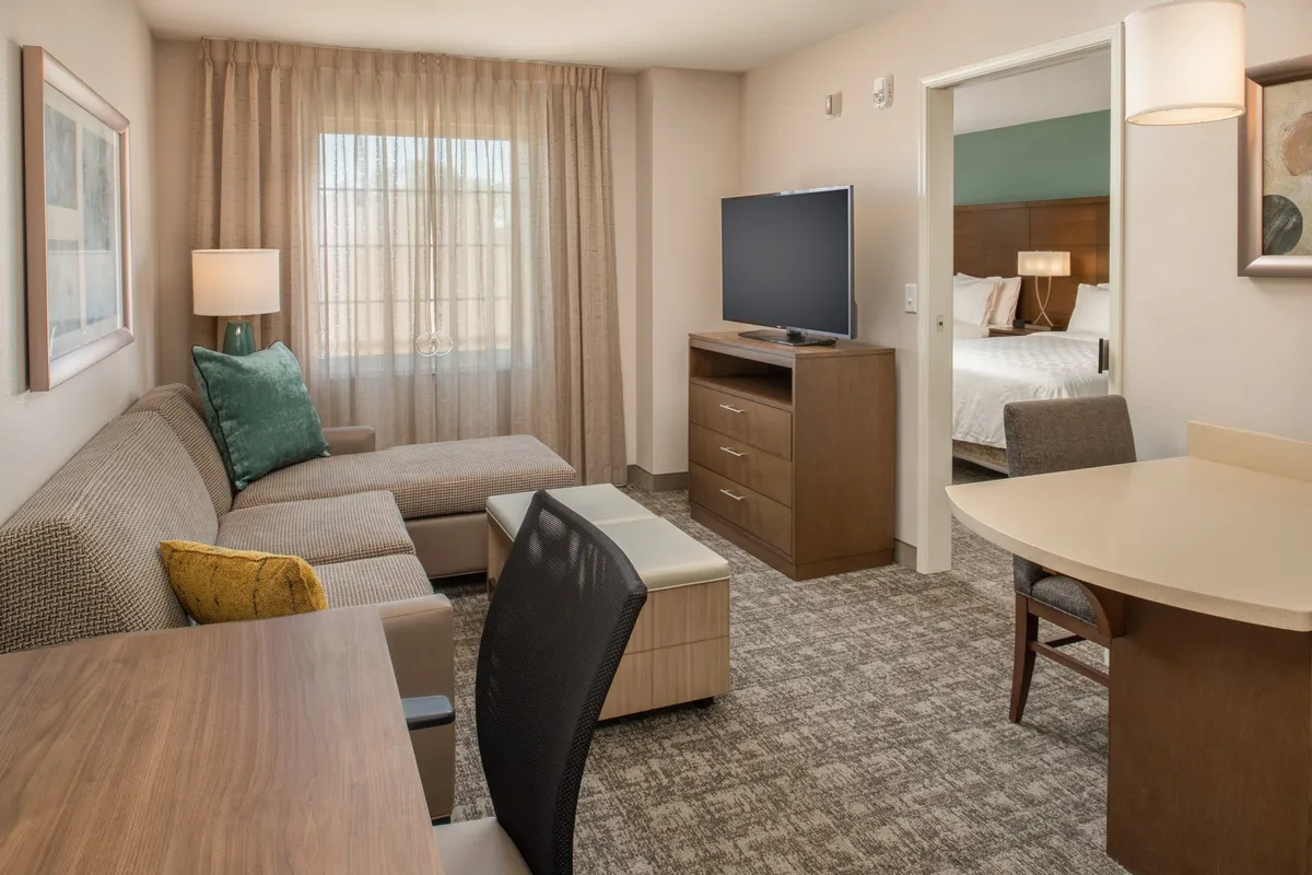 The hero image showcases a cozy, modern living area with a plush sofa facing a large TV, a dining table perfect for meals or remote work, and a glimpse into the peaceful bedroom. Natural light streams through inviting curtains, creating a warm and welcoming atmosphere ideal for relaxation or productivity.