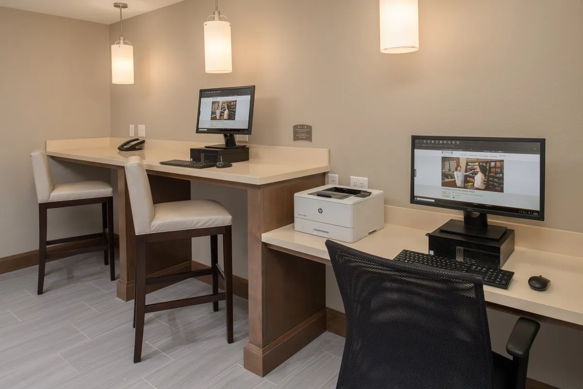 An inviting workspace with computers, a printer, and bar-style seating. The setup is modern and conducive to productivity, with ample light and accessible technology for working or connecting online.