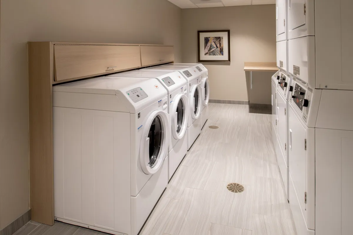 A practical laundry room equipped with multiple washing machines, dryers, counter space for folding, and framed artwork on the wall. The organized environment suggests ease and convenience for guests planning longer stays.